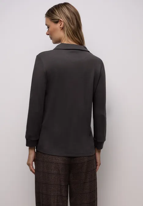 Frau in dunklem Langarmshirt und karierten Hosen steht mit dem Rücken zur Kamera vor hellem Hintergrund.